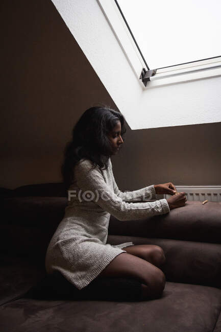 Side view of calm young Indian woman in casual dress resting on comfortable sofa near window in modern attic room while rolling up marijuana cigaret — Stock Photo