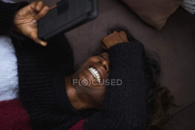 Зверху весела молода етнічна леді в повсякденному теплому светрі, що розслабляється на зручному м'якому дивані і посміхається при використанні смартфона — стокове фото