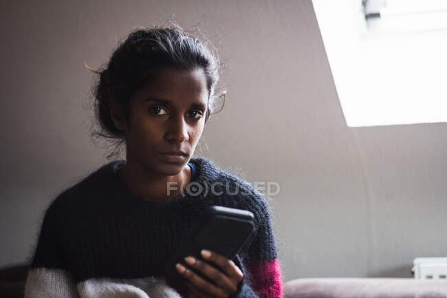 Спокійна молода етнічна леді з темним волоссям у випадковому теплому светрі переглядає смартфон, відпочиваючи на зручному дивані в світлій квартирі — стокове фото