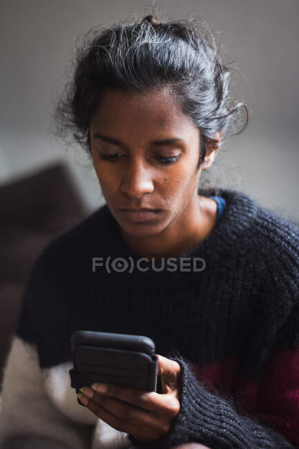 Calm young ethnic lady with dark hair in casual warm sweater browsing smartphone while resting on comfortable couch in light apartment — Stock Photo