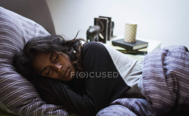 Вид сбоку отдыхающей индийской женщины в сонной одежде, мирно отдыхающей на уютной кровати на мягкой подушке и одеяле в светлой спальне — стоковое фото