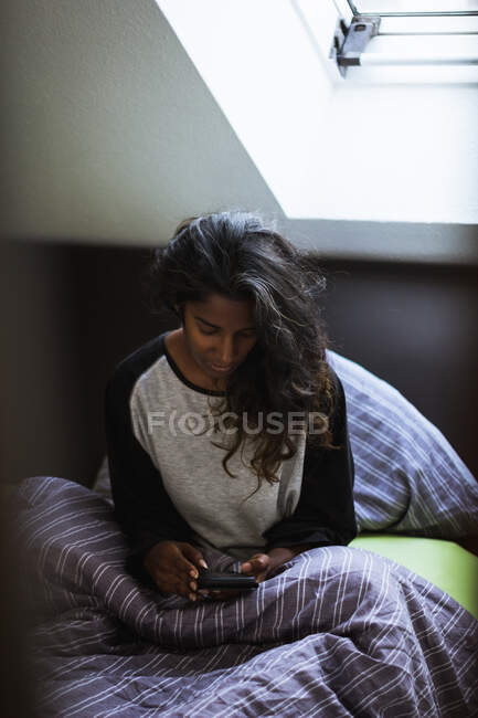 Vista lateral da mulher indiana pensativa em roupas de dormir sentado na cama acolhedora no travesseiro macio e cobertor usando telefone celular no quarto leve — Fotografia de Stock