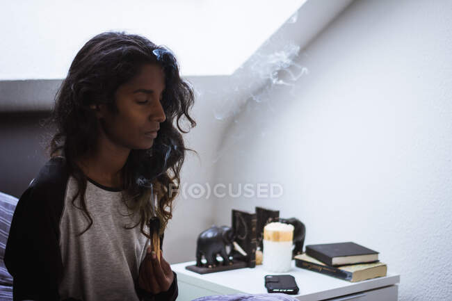 Vista lateral da fêmea indiana consciente com longos cabelos escuros ondulados em roupas de dormir descansando na cama enquanto desfruta de queimar paus de incenso no apartamento moderno — Fotografia de Stock