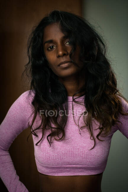 Attraktive junge ethnische Dame mit langen welligen Haaren in trendiger pinkfarbener Kleidung steht in modernem Raum mit den Händen auf der Taille und blickt nachdenklich in die Kamera — Stockfoto