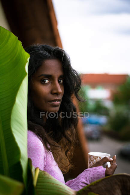 Schöne ethnische Frau mit langen dunklen Haaren trägt rosa Freizeitkleidung und hält eine Tasse mit frischem Heißgetränk in den Händen, während sie auf dem Balkon in der Nähe des pflanzengrünen Blattes steht und in die Kamera schaut — Stockfoto