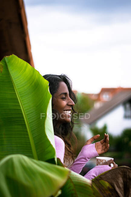 Mujer étnica joven positiva con el pelo largo y oscuro usando ropa casual rosa sosteniendo la taza de bebida caliente fresca en las manos mientras está de pie en el balcón cerca de la hoja verde de la planta mirando hacia otro lado - foto de stock