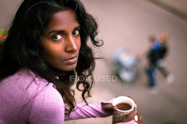 Сверху вид молодой этнической женщины с длинными темными волосами в розовой повседневной одежде, держащей чашку горячего напитка в руках, стоя на балконе и глядя в камеру — стоковое фото