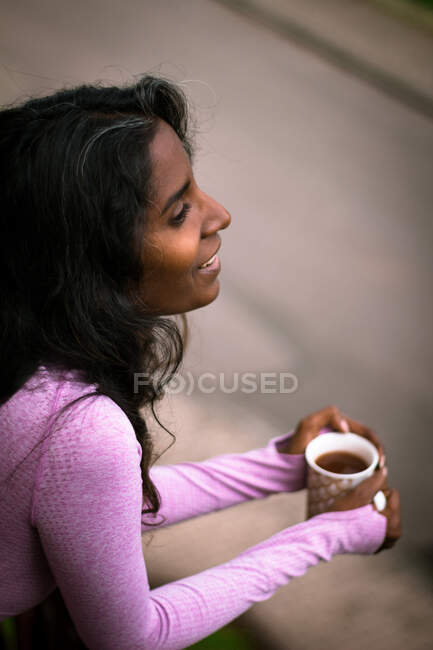 Сверху вид положительной молодой этнической леди с длинными темными волосами в розовой повседневной одежде, держащей чашку свежего горячего напитка в руках, стоя на балконе и глядя в сторону — стоковое фото