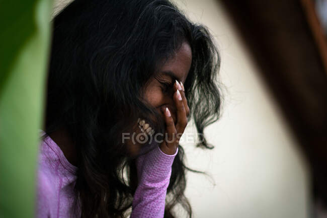 Вид сбоку смеющейся молодой этнической леди с длинными черными волосами, покрывающими лицо ладонью, веселящейся на террасе — стоковое фото