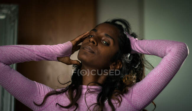 Sensual joven mujer étnica en ropa rosa informal de pie en el apartamento moderno y tocando el pelo largo y ondulado negro mientras mira a la cámara - foto de stock