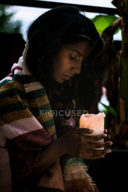 Concentrated young ethnic female shaman in warm poncho holding candle while performing spiritual ceremony in evening — Stock Photo