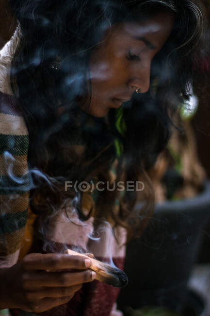 Mujer espiritual joven étnica con los ojos cerrados en ropa tradicional sosteniendo incienso de salvia ardiente con humo y meditando al aire libre - foto de stock