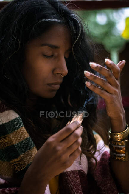 Jeune femme ethnique spirituelle les yeux fermés dans des vêtements traditionnels tenant de l'encens de sauge brûlant avec de la fumée et méditant à l'extérieur — Photo de stock