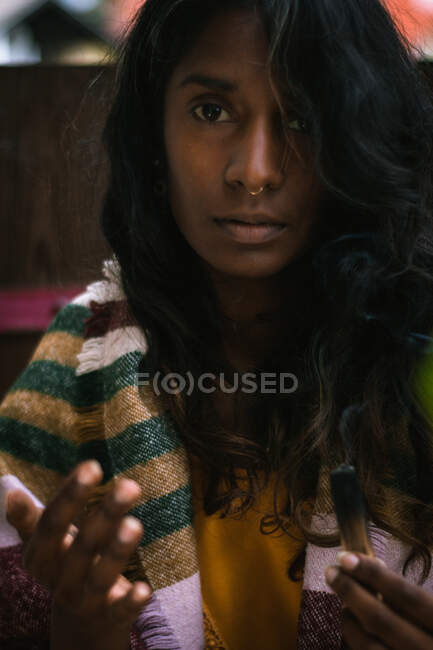 Spirituale giovane femmina etnica guardando la macchina fotografica in abiti tradizionali che tengono bruciare incenso salvia con fumo e meditando all'aperto — Foto stock