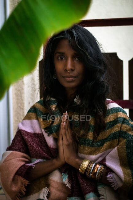 Joven mujer espiritual étnica en ropa tradicional manteniendo las manos en gesto namaste y mirando a la cámara - foto de stock