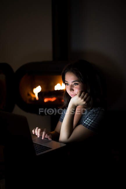 Жінка в повсякденній сорочці сидить у темній затишній кімнаті біля каміну і читає книгу на нетбуці, відпочиваючи вночі — стокове фото