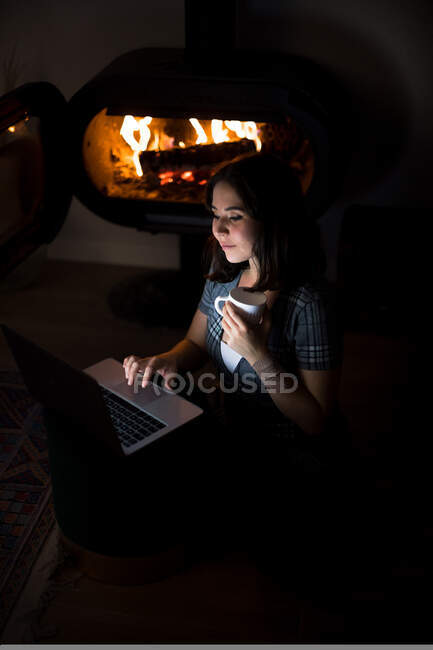 Сверху содержание женщины сидит на полу и согревается с чашкой горячего напитка во время использования нетбука и охлаждения в выходные дни дома — стоковое фото