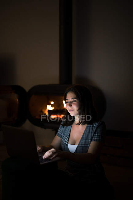 Donna pensierosa in maglietta casual seduta in camera buia accogliente vicino al camino e leggere libro su netbook mentre riposa durante la notte — Foto stock