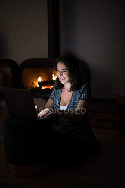 Offenes Weibchen in lässigem T-Shirt sitzt in dunklem, gemütlichem Raum am Kamin und liest Buch auf Netbook, während es sich nachts ausruht — Stockfoto