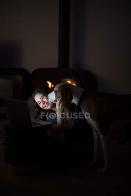 De cima feliz fêmea sentada no chão na sala de estar escura com lareira e abraçando Harrier enquanto assistia vídeo no laptop juntos — Fotografia de Stock