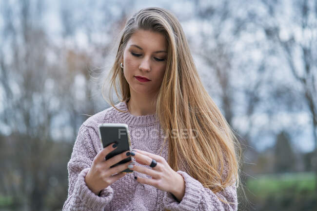 Jeune femme détendue en pull décontracté regardant les médias sociaux sur smartphone et souriant assis sur un banc en pierre dans la rue — Photo de stock