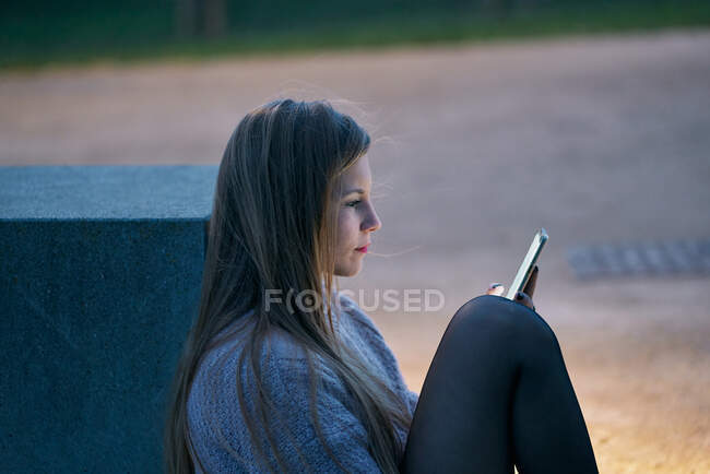 Mujer joven feliz en jersey casual viendo las redes sociales en el teléfono inteligente y sonriendo mientras se sienta en el banco de piedra en la calle - foto de stock