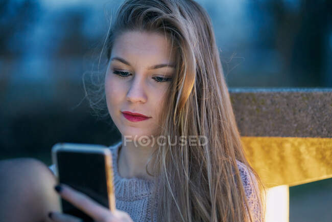 Chica joven que utiliza su teléfono celular por la noche apoyada en un banco - foto de stock