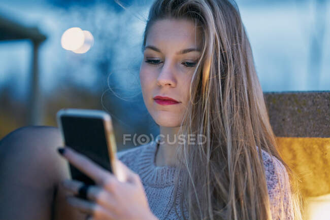 Молодая девушка, которая использует свой мобильный телефон ночью опираясь на скамейку — стоковое фото
