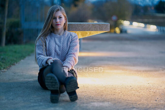 Серьёзная блондинка в свитере и юбке смотрит в камеру, сидя на бетонной дорожке и вытягивая скрещенные ноги к камере — стоковое фото