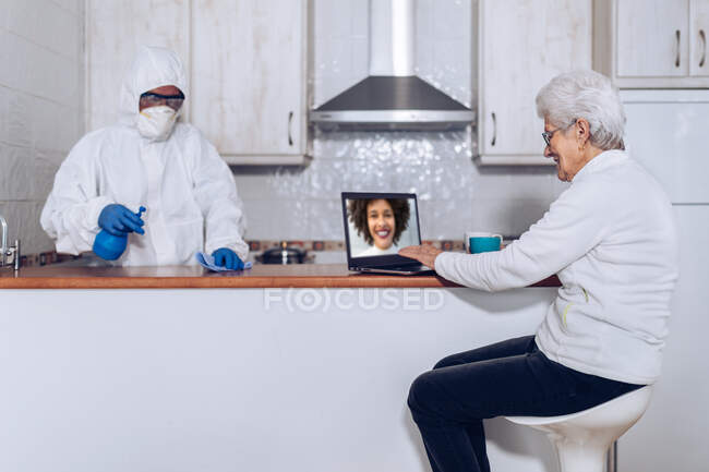 Mujer mayor sentada en el mostrador de la cocina y comunicándose con sus familiares durante una videollamada a través de computadora portátil mientras trabaja en el hogar con uniforme protector y enmascara muebles desinfectantes durante la cuarentena del coronavirus - foto de stock