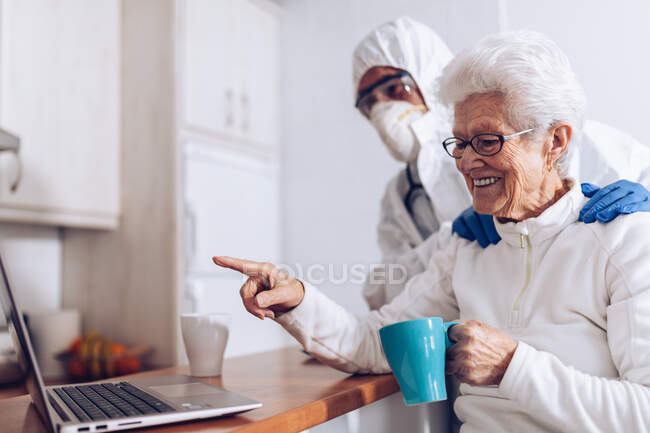 Alegre vieja mujer bebiendo té y charlando con un amigo durante la videollamada a través de un ordenador portátil, mientras que el especialista en el cuidado del hogar en traje de protección y máscara de pie cerca durante el bloqueo coronavirus - foto de stock