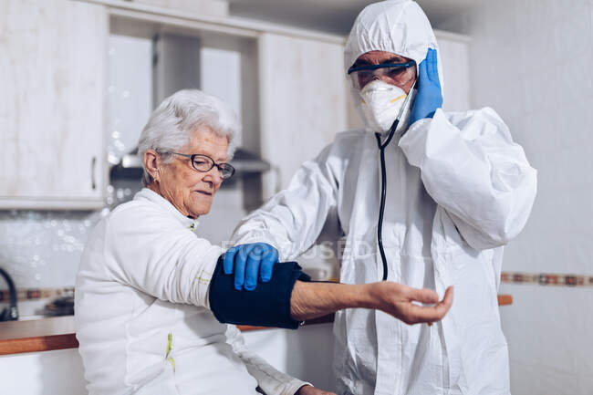 Assistente de cuidados domiciliares profissional em uniforme de proteção e máscara examinando a mulher idosa e verificando a pressão arterial ao visitar o paciente em casa durante o surto de coronavírus — Fotografia de Stock