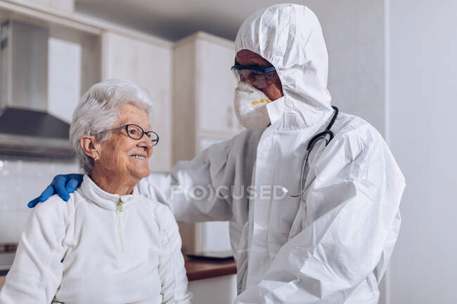 Неузнаваемый мужчина-сиделка в защитном костюме и маске со стетоскопом, который поддерживает и разговаривает с улыбчивой пожилой женщиной во время посещения пациента дома во время блокировки коронавируса — стоковое фото