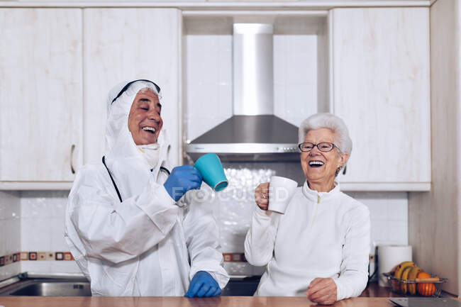 Happy gray haired female and male caregiver in protective uniform and gloves drinking tea and having fun while standing at kitchen counter during coronavirus lockdown — Stock Photo