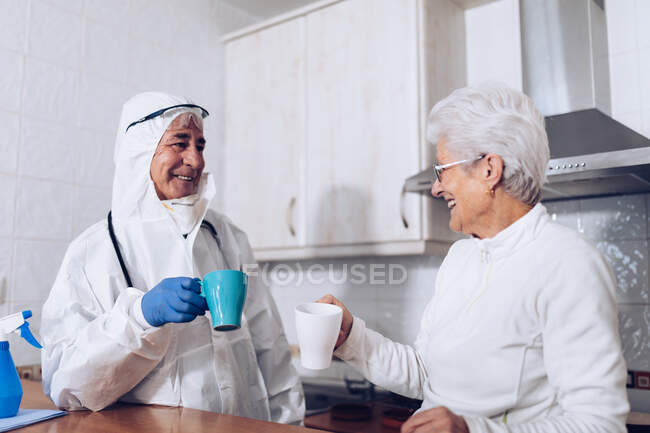 Mujer mayor alegre con cuidador bebiendo té en la cocina durante la cuarentena - foto de stock