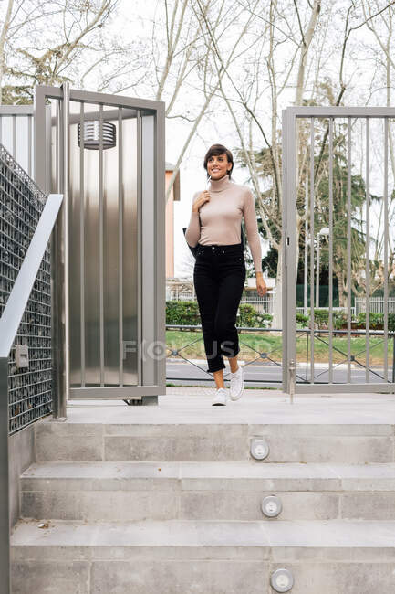 Adulto feliz morena feminina em roupa casual e cachecol cinza pisando em escadas de pavimento de concreto no dia ensolarado e olhando para longe — Fotografia de Stock