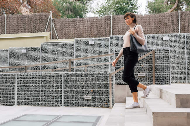 Доросла щаслива брюнетка-жінка в повсякденному вбранні і сірий шарф ступає на бетонні сходи на тротуарі в сонячний день і дивиться в сторону — стокове фото