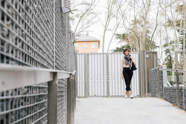 Доросла щаслива брюнетка жінка в повсякденному вбранні і сірий шарф ступає на бетонну тротуарну плиту за металевими воротами в сонячний день і дивиться на камеру — стокове фото