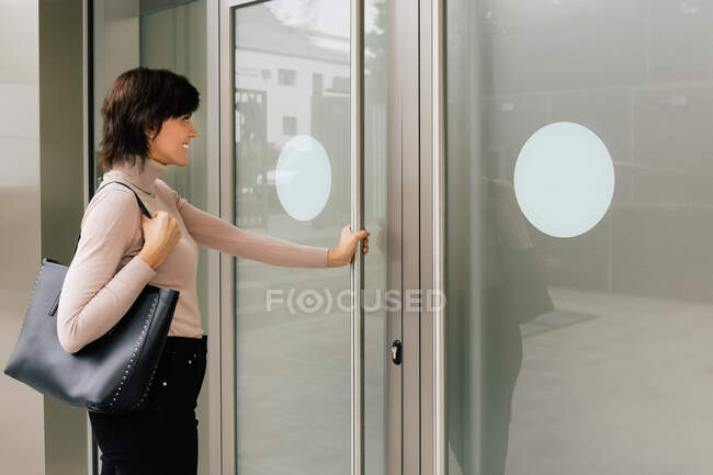 Side view of cheerful brunette female with short hair in casual outfit and with black handbag taking metal door handle while standing outside in sunlight — Stock Photo