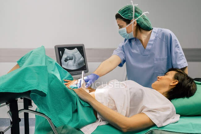 Médica feminina em máscara estéril e luva azul usando scanner de ultra-som enquanto examina a mulher grávida alegre e olhando para a tela do computador no hospital — Fotografia de Stock