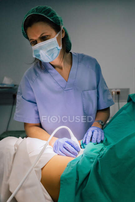 Médecin féminin dans un masque stérile et un gant bleu à l'aide d'un scanner à ultrasons tout en examinant la femme enceinte et en regardant l'écran d'ordinateur à l'hôpital — Photo de stock