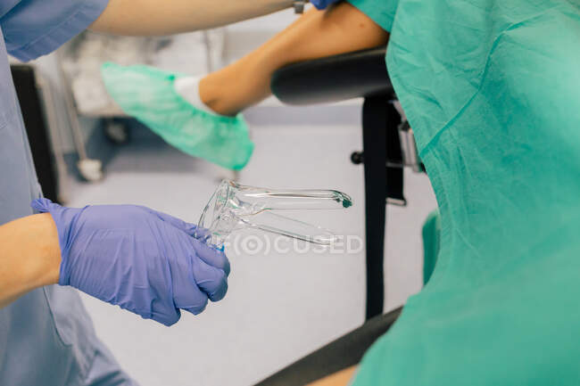 Médica sem rosto em luvas azuis e uniforme com espelho ginecológico de plástico examinando paciente anônimo em capas de sapato deitado na cadeira na clínica de fertilidade — Fotografia de Stock