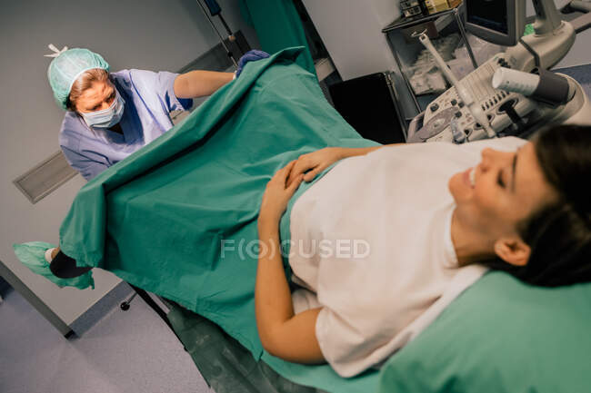 Doctora en uniforme azul y máscara estéril examinando paciente sonriente en silla ginecológica en clínica de fertilidad - foto de stock