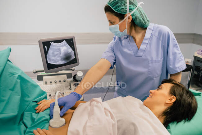 Médica feminina em máscara estéril e luva azul usando scanner de ultra-som enquanto examina a mulher grávida alegre no hospital — Fotografia de Stock