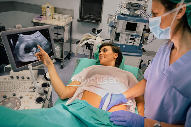 Médecin féminin dans un masque stérile et un gant bleu à l'aide d'un échographe tout en examinant gaie femme enceinte pointant vers l'écran à l'hôpital — Photo de stock