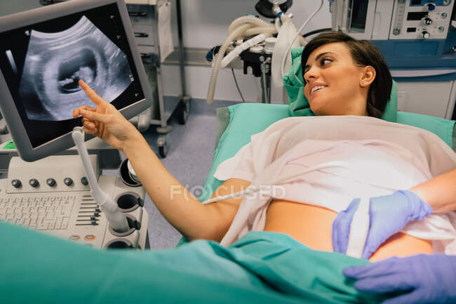 Médecin méconnaissable dans des gants bleus à l'aide d'un échographe tout en examinant gaie femme enceinte pointant vers l'écran à l'hôpital — Photo de stock