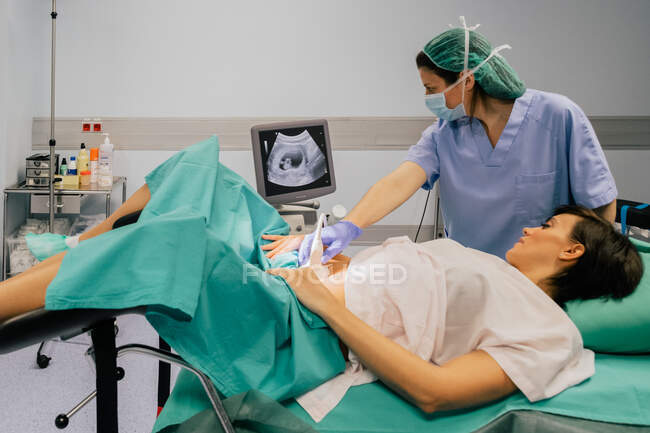 Doctora en máscara estéril y guante azul usando escáner de ultrasonido mientras examina a una mujer embarazada alegre en el hospital - foto de stock