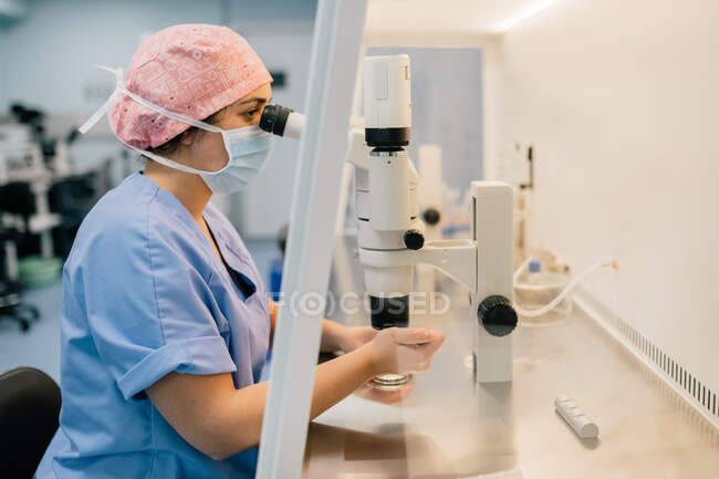 Von oben Arzt in medizinischer Maske und gleichmäßiger Injektion der Eizelle auf Petrischale und Untersuchung der Zelle unter dem Mikroskop im Labor der modernen Fruchtbarkeitsklinik — Stockfoto