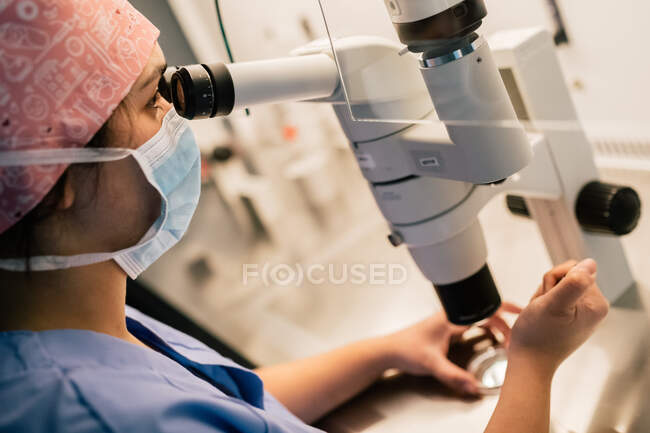 From above doctor in medical mask and uniform injecting ovum on Petri dish and examining cell through microscope in laboratory of modern fertility clinic — Stock Photo