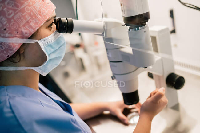 From above doctor in medical mask and uniform injecting ovum on Petri dish and examining cell through microscope in laboratory of modern fertility clinic — Stock Photo
